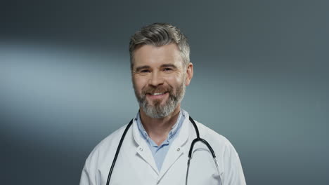 Portrait-shot-of-the-Caucasian-handsome-gray-haired-male-doctor-in-the-white-gown-and-with-stethoscope-on-his-neck-smiling-cheerfully-to-the-camera.-Close-up.